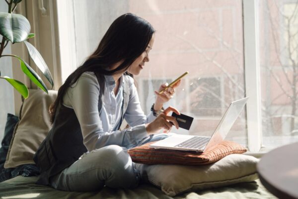 Woman with long dark hair sitting on a bed or couch next to a window looking at her phone to illustrate Holiday Deals Are Here. Don't Let Debt Follow