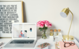 A macbook, phone, coloured notebooks, a gold lamp, and a vase of pink roses on a white desk