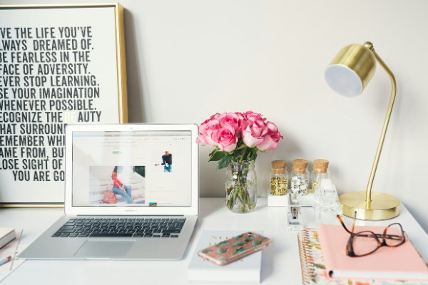 A macbook, phone, coloured notebooks, a gold lamp, and a vase of pink roses on a white desk